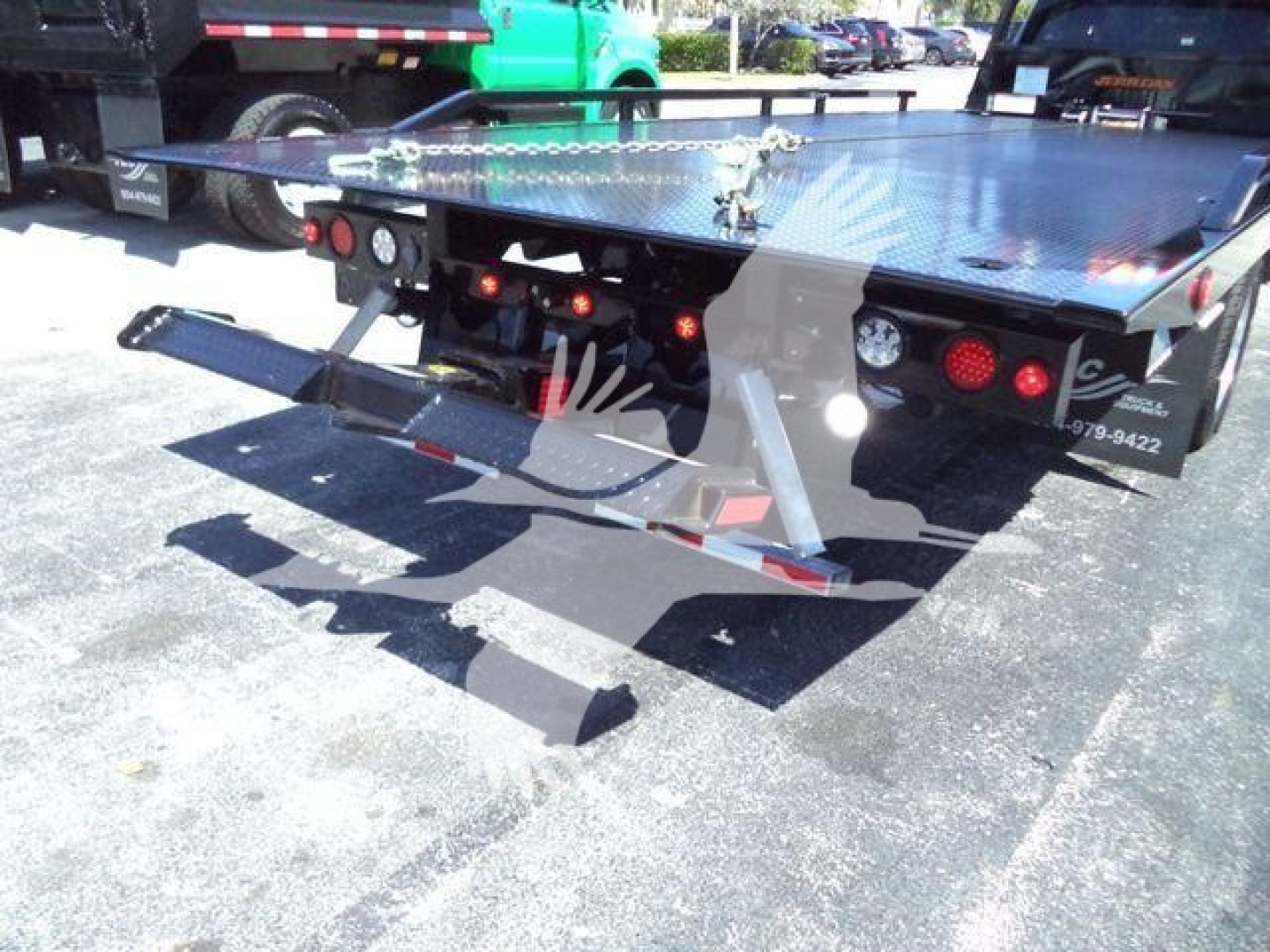 Truck bed setup with a diamond plate platform and hydraulic rollback equipment; rear lights and safety chains visible. Some text on the rear panel shows numbers and contact info.