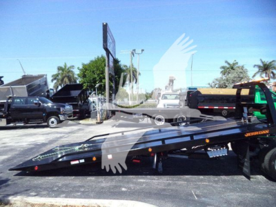 Tow truck or rollback equipment with a folded flatbed and a Ram 5500 in the lot. Visible text includes 'JerrDan' on the bed, and vehicle in foreground. Scene shows other trucks and palm trees.