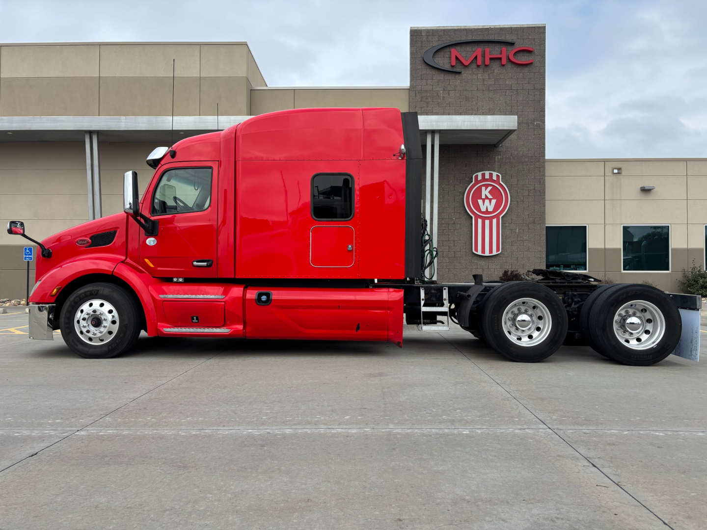 Peterbilt 579 tractor unit in red side view, parked in front of a building with MHC KW logo, two-axle sleeper cab and extended chassis.