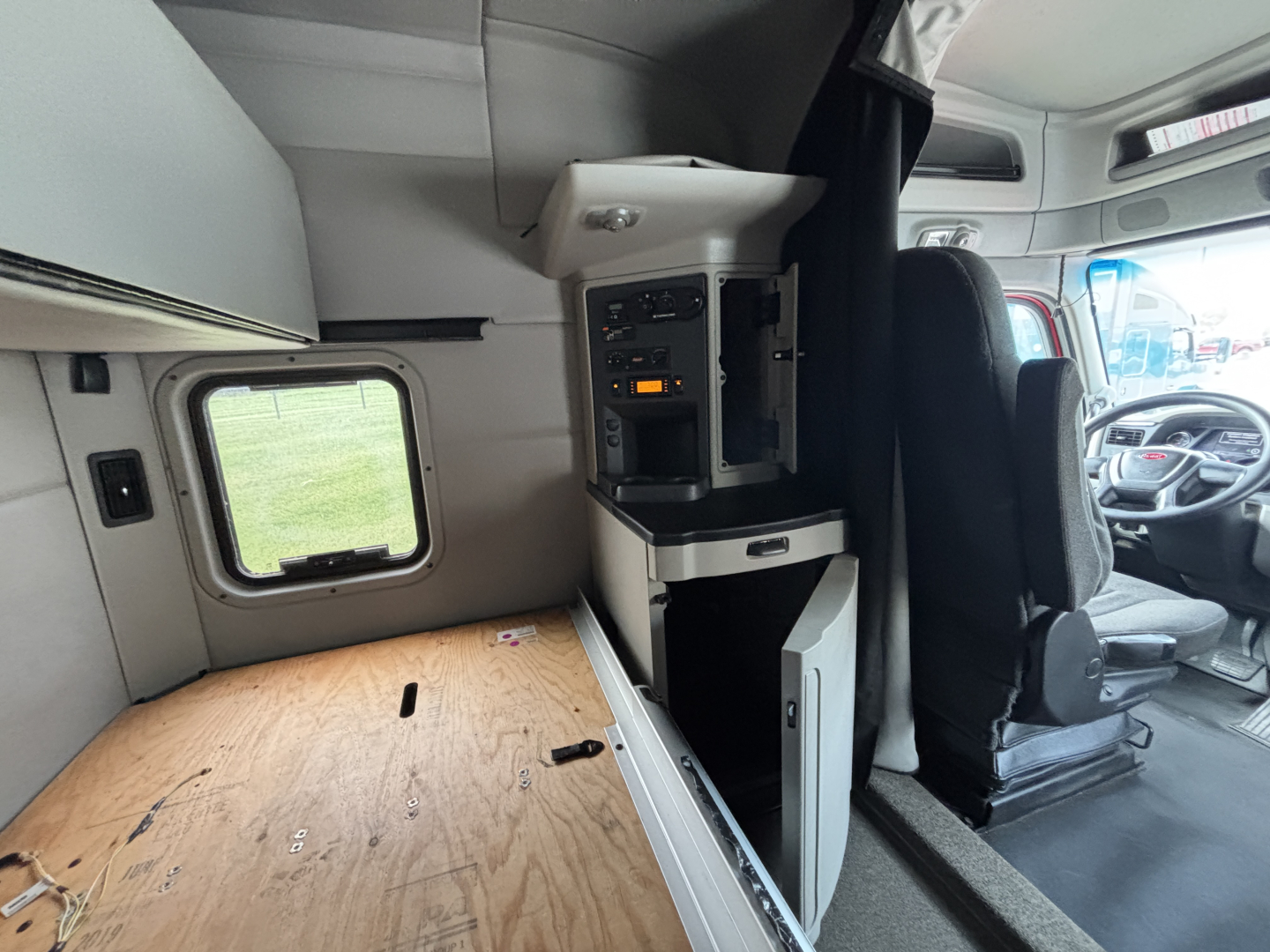 Peterbilt 579 cab interior showing driver seat with dashboard and overhead storage, wood plank floor area and passenger side cab access.