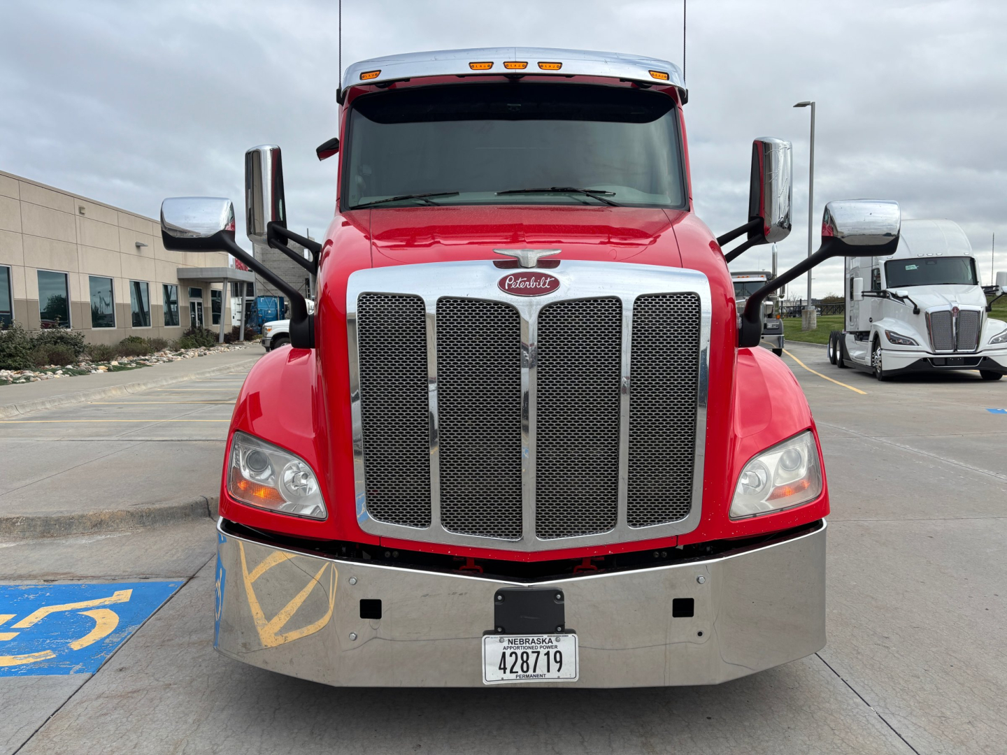 Peterbilt 579 truck front view showing large chrome grille, red hood, and bumper with license plate; mirrors extend from sides; headlights visible.
