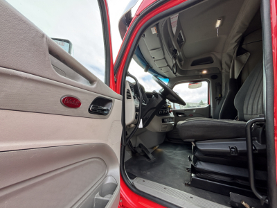 Peterbilt truck interior with open door showing driver seat, steering wheel, dashboard; red exterior; door panel has Peterbilt emblem.