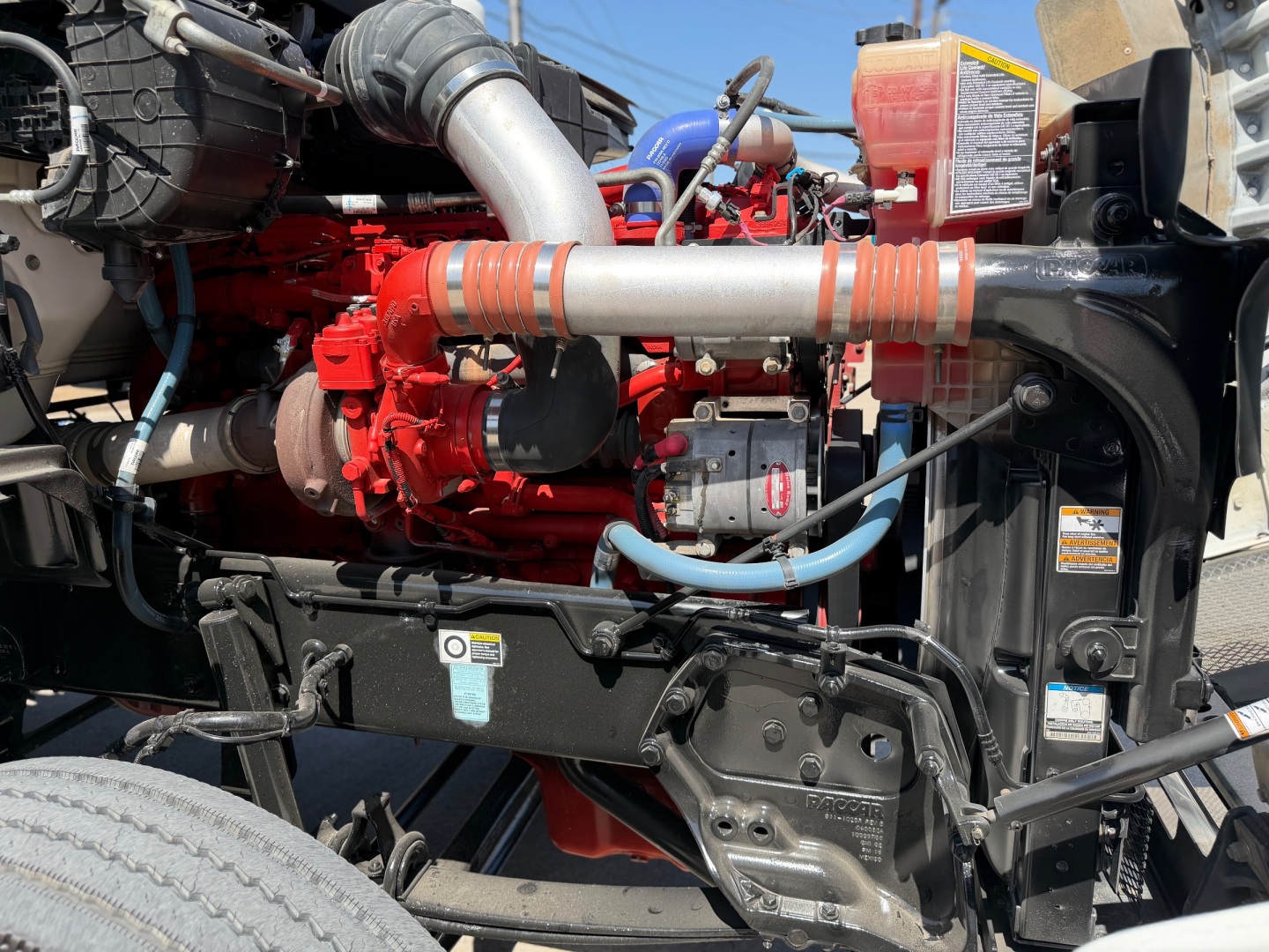 Kenworth T880 engine bay showing red engine, silver hoses, blue wiring, and black front frame with warning labels.