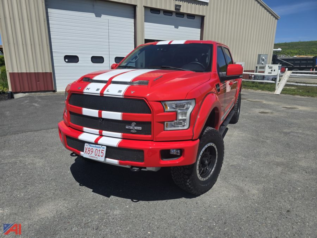 Front view of a red 2016 Ford Shelby F150 crew cab pickup with white racing stripes, black grille, raised stance, on a parking lot in front of a beige building.