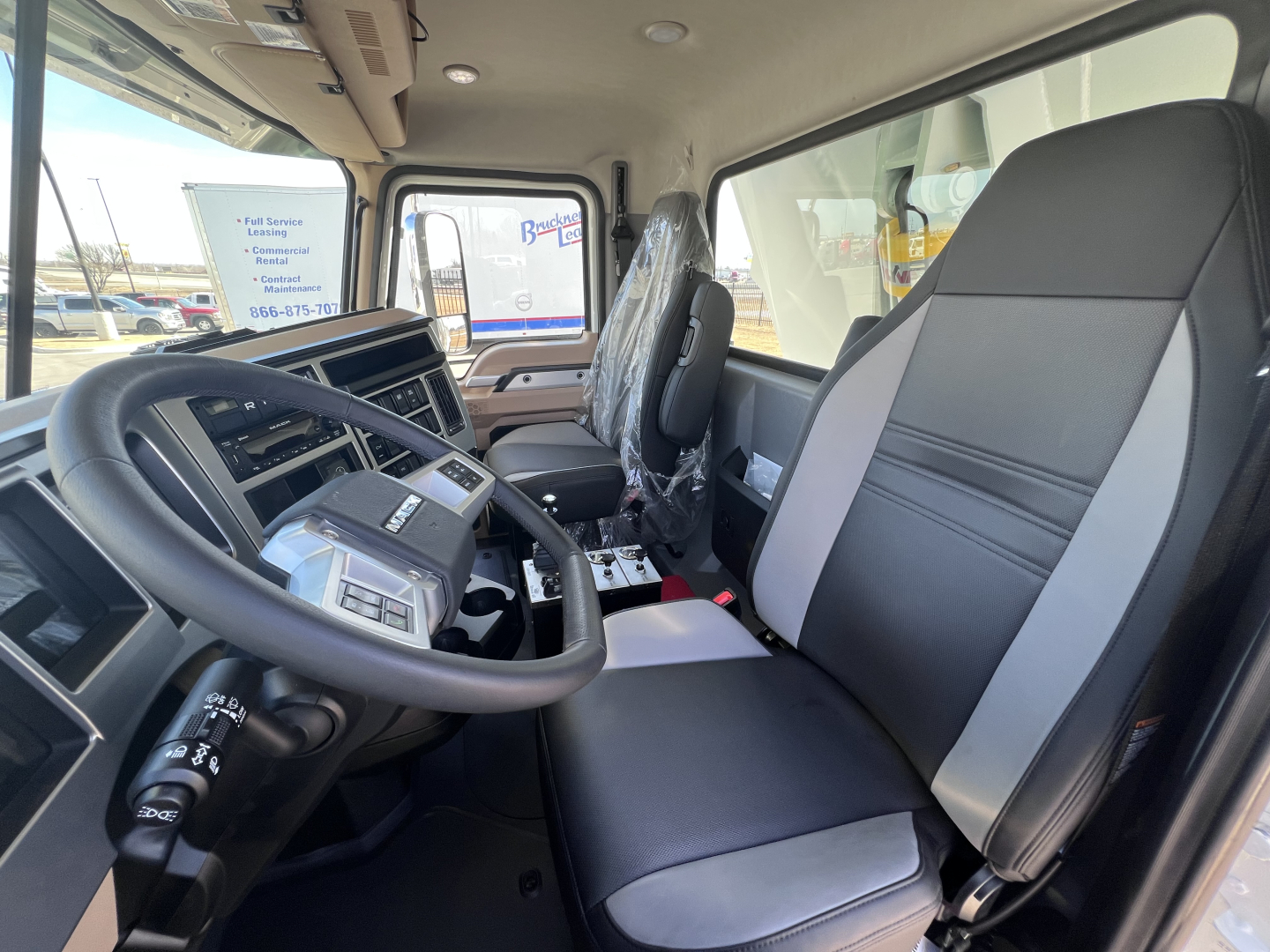 Cab interior of a 2026 Mack GR64F dump truck showing steering wheel, dashboard, front seats with plastic-wrapped passenger seat, and center console.