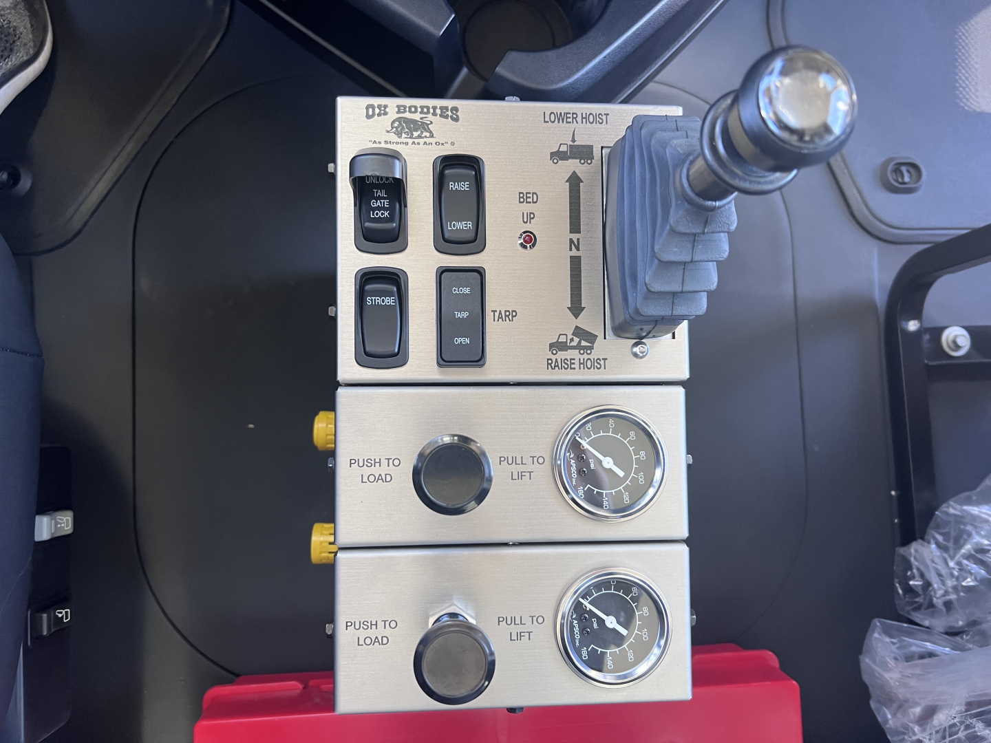 Mack GR64F truck control panel with multiple switches labeled THEZ? LOWER HOIST, BED UP, TARP, PULL TO LIFT, PUSH TO LOAD, and two round gauges; joystick-like lever on right; metallic panels.