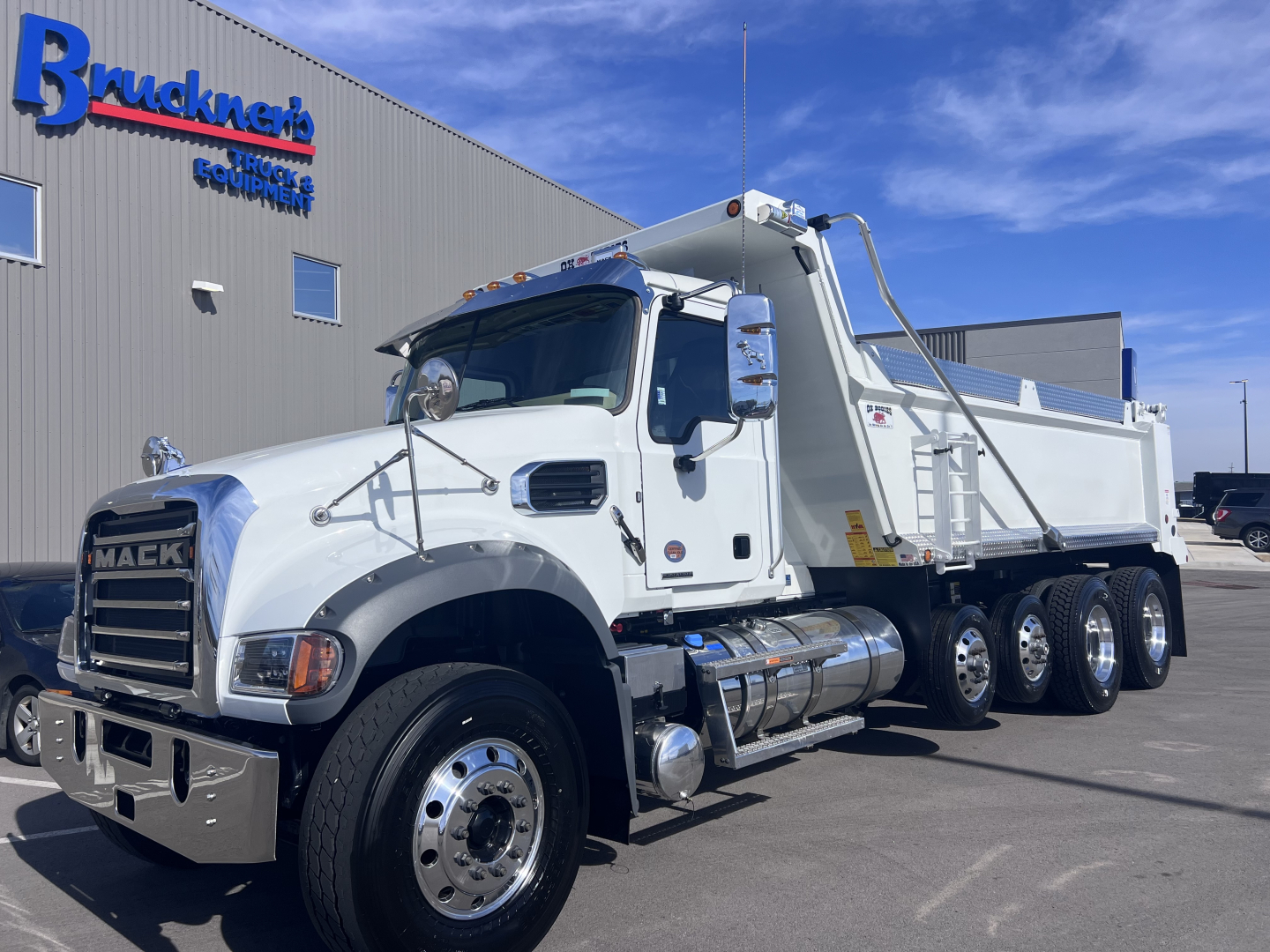Dump truck showing Mack GR64F with white cab and dump bed, chrome front bumper, dual rear axles, side steps, and company building in background.