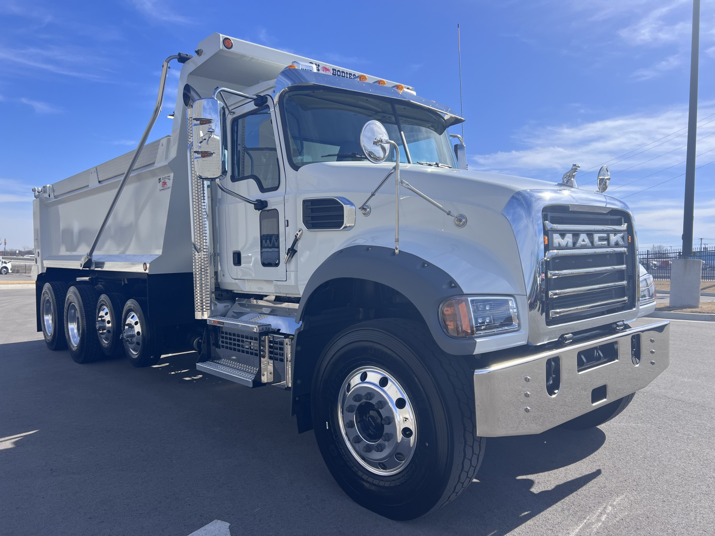 Mack GR64F dump truck front-left view showing chrome grille and dual rear axles with aluminum dump body.