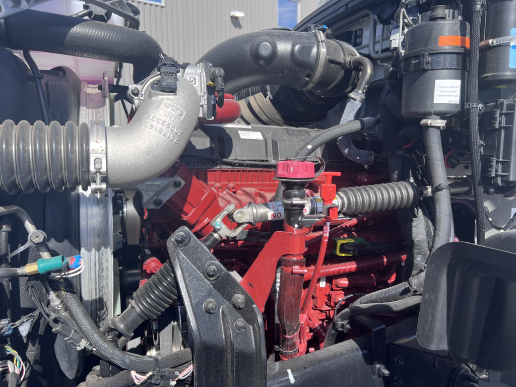 Mack GR64F engine bay showing red Cummins/Detroit engine block with turbo piping and hoses in view.