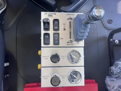 Mack GR64F truck control panel with multiple switches labeled THEZ? LOWER HOIST, BED UP, TARP, PULL TO LIFT, PUSH TO LOAD, and two round gauges; joystick-like lever on right; metallic panels.