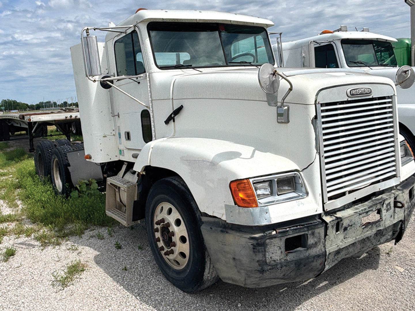 Freightliner FLD112 conventional truck front-left view showing cab, grille, and bumper with worn lower fascia, DOT mirrors, and mid-2000s styling.