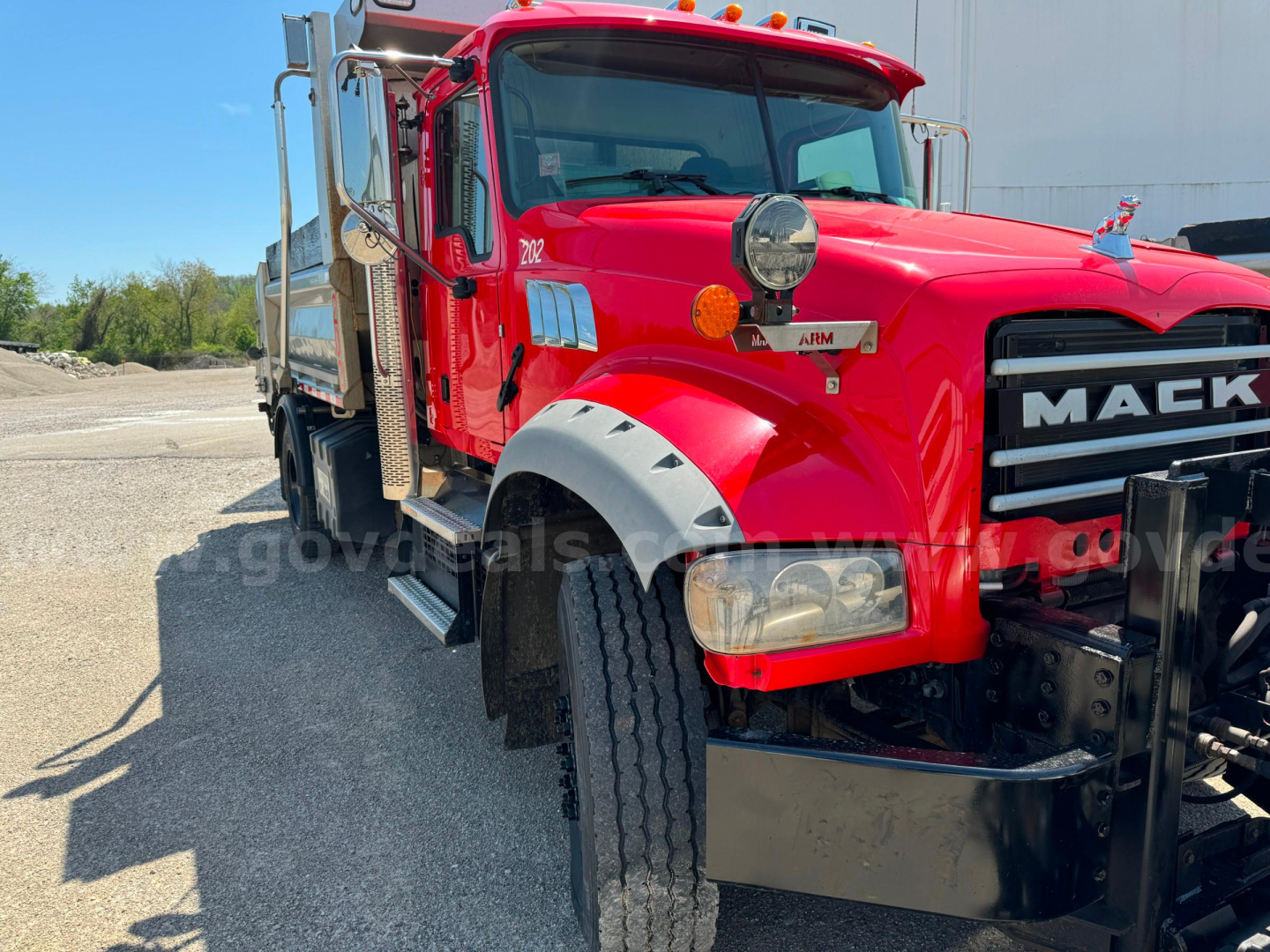 Mack Granite medium-duty truck front-left view showing red cab, large round auxiliary light, chrome grille with MACK letters, side mirrors, and plow/utility equipment on the bed.