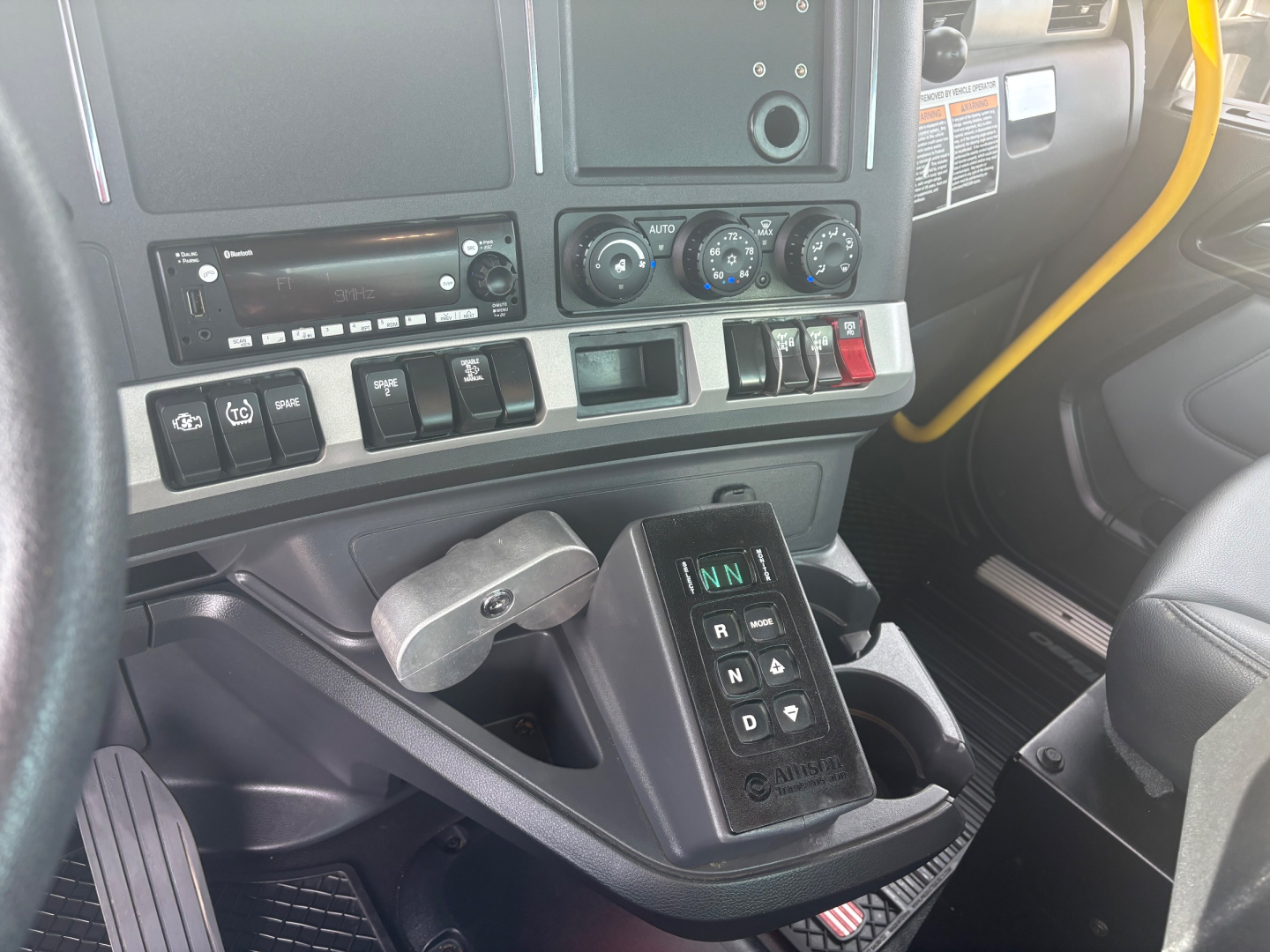 Interior view of a Kenworth T880 cabin showing gear selector, dashboard controls, and climate dials. Windows, steering column, and part of passenger seat visible.