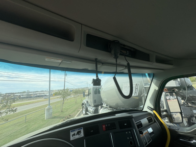 Truck cab interior overhead view showing upper dash area, CB microphone, and sun visor with a concrete mixer in the background.