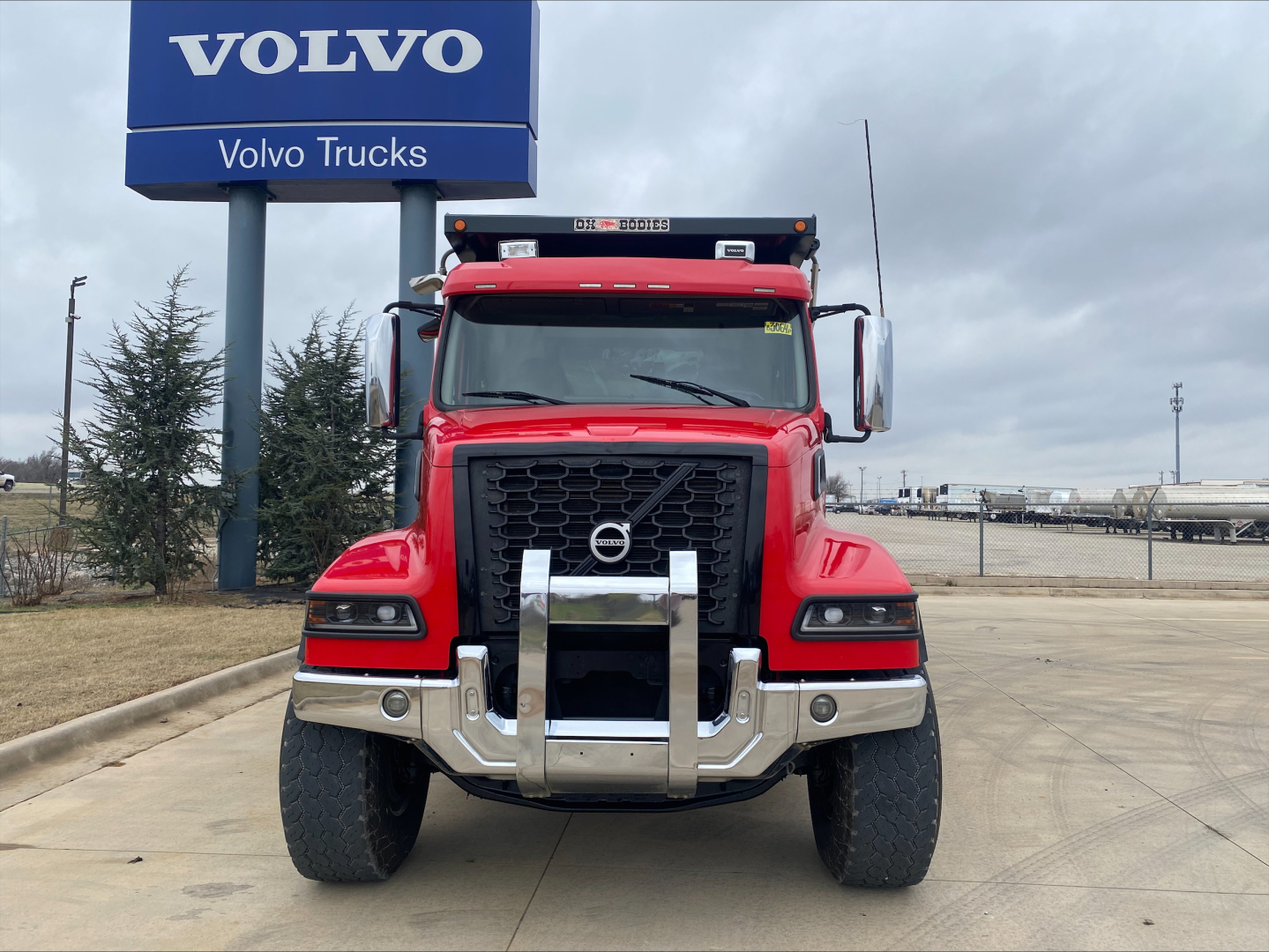 Volvo VHD truck front view in red with large chrome grille guard and bumper, Volvo sign in background, no visible text on truck aside from model features.