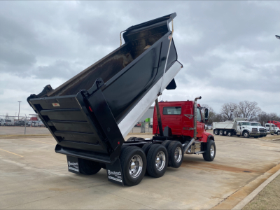Dump truck with raised black dump bed, red cab, three rear axles, in a lot.