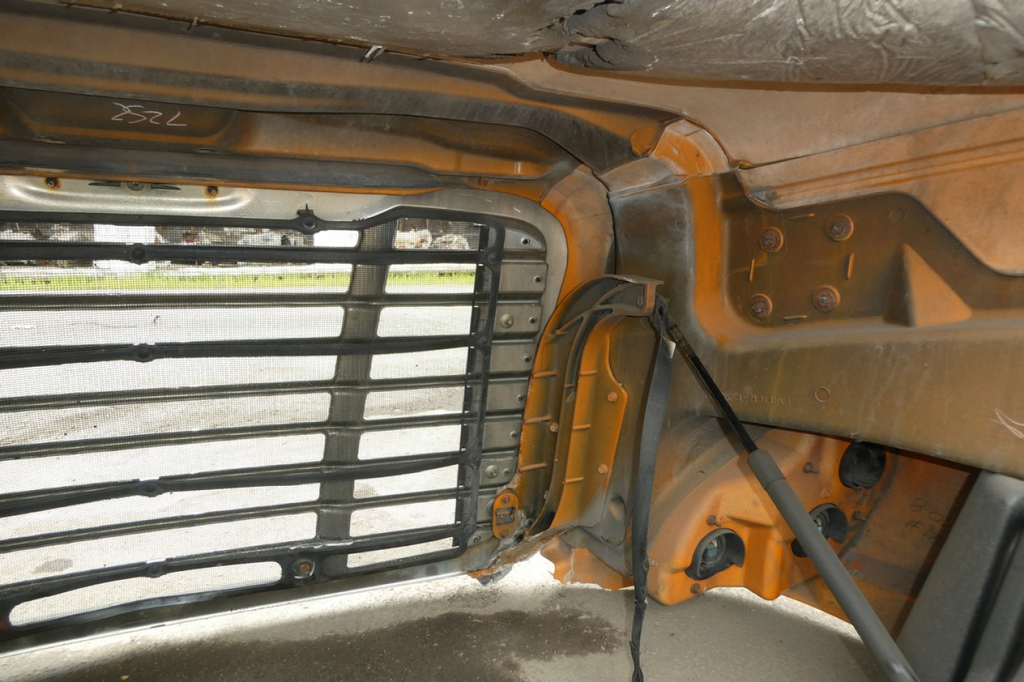 Cab interior near side window with metal louvered grille and orange-painted frame, hydraulic lines, and wiring harnesses visible.