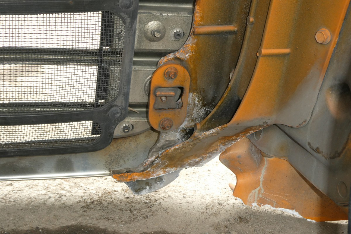 Front left corner of a Freightliner Cascadia 125 cab showing chipped orange paint, rust-looking residue near bumper, grille area, and hinge/strap nearby.