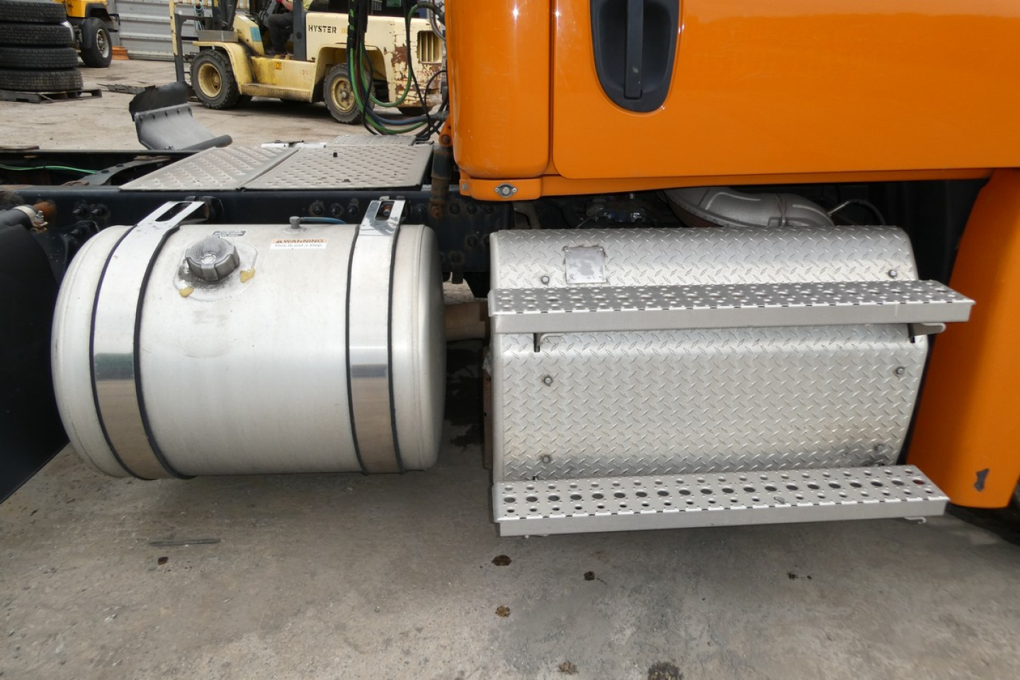 Truck side view showing aluminum cylindrical fuel tank and diamond-plate step on Freightliner Cascadia 125 cab; orange exterior visible above; metallic components with bolts.