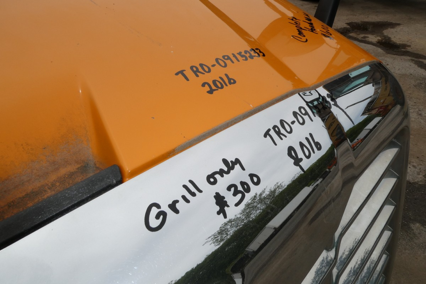 Truck hood with handwritten notes: 'Grill only $300' and '2016' near windshield, and other markings on orange cab and chrome mirror; reflective surface shows partial windshield text.