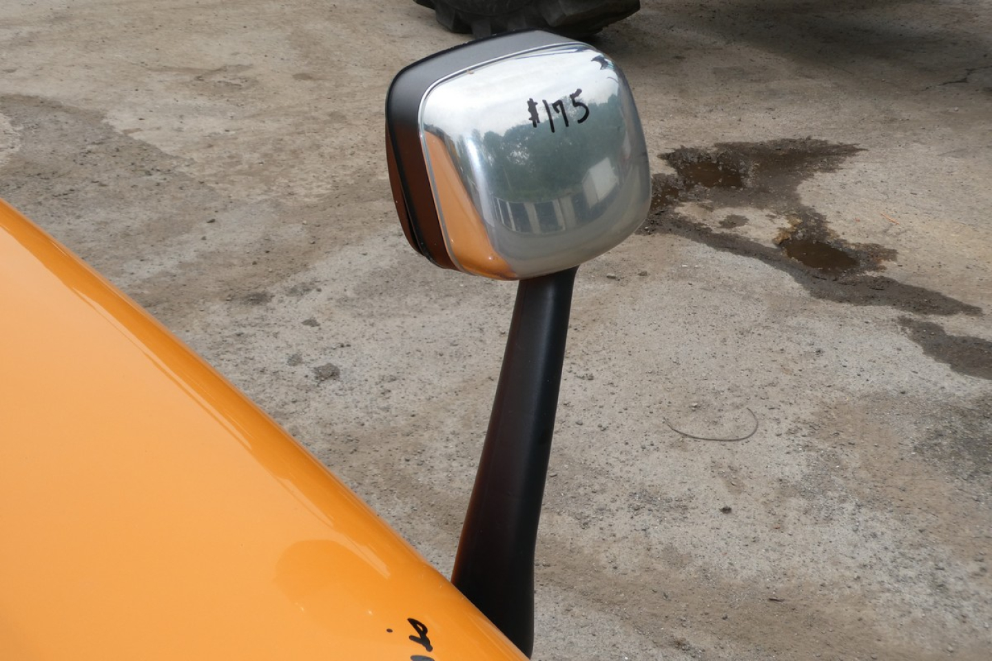 Exterior close-up of a Freightliner Cascadia 125 mirror assembly with orange cab and black mounting, showing part of the door and reflective mirror with '#115' marking.