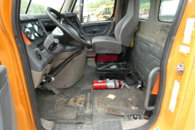 Cab interior of a 2017 Freightliner Cascadia 125 Daycab showing driver seat, steering wheel, dashboard, and floor area with a fire extinguisher and scattered debris.