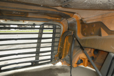 Cab interior near side window with metal louvered grille and orange-painted frame, hydraulic lines, and wiring harnesses visible.