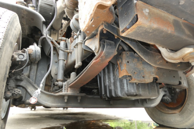Truck undercarriage showing front axle, suspension, and engine bay components with frame rails and hoses.