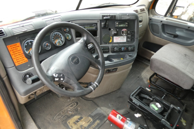 Interior of a Freightliner Cascadia cab: steering wheel, dashboard with gauges, radio, and control panel. Passenger seat and floor mat visible.