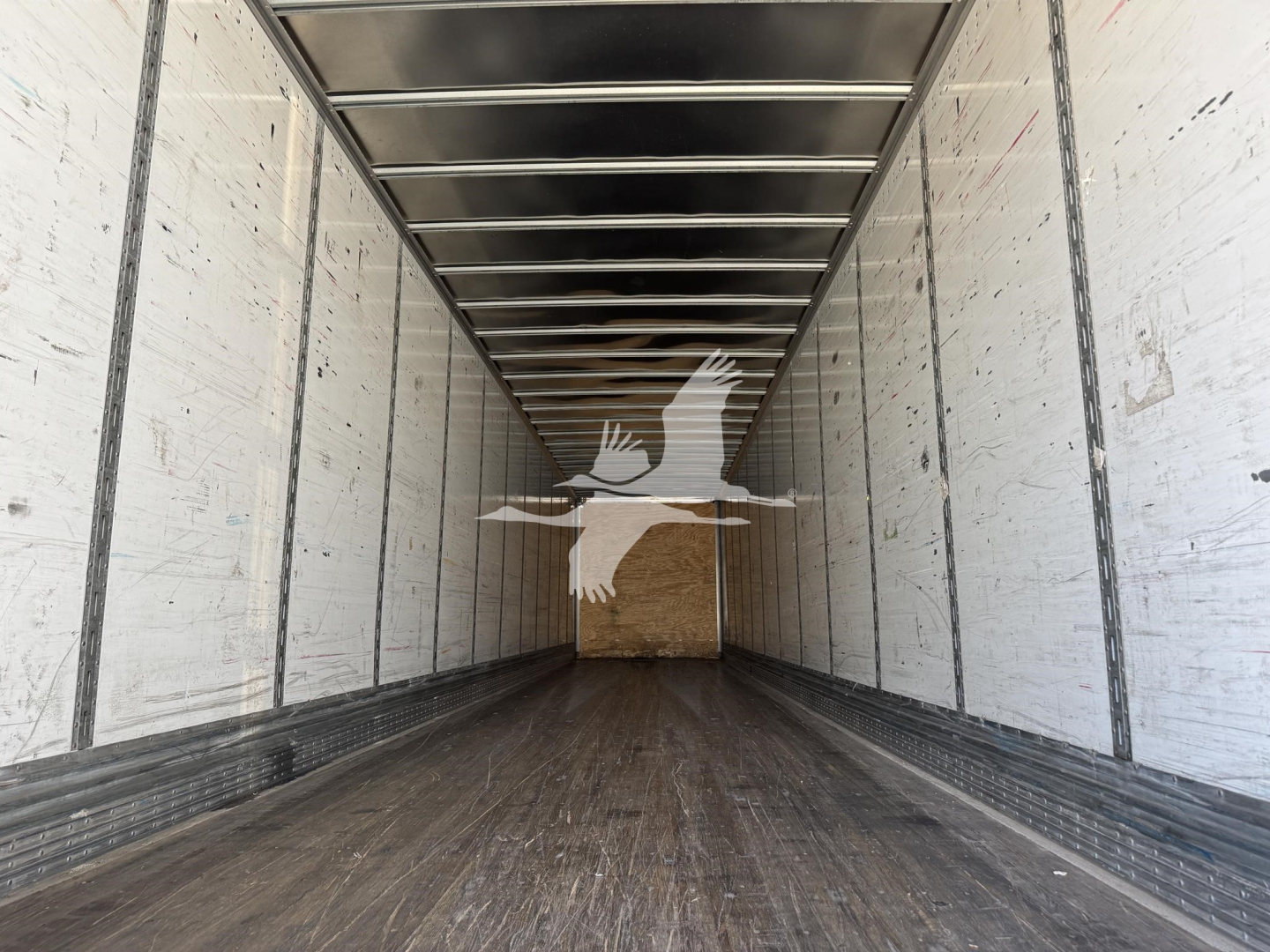 Interior of a trailer cargo space showing empty wooden floor, sidewalls with riveted panels, and overhead support beams.