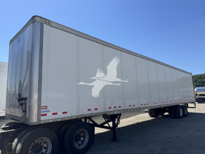 Truck trailer side view showing white Vanguard logo on gray trailer with riveted panels and dual rear axles.