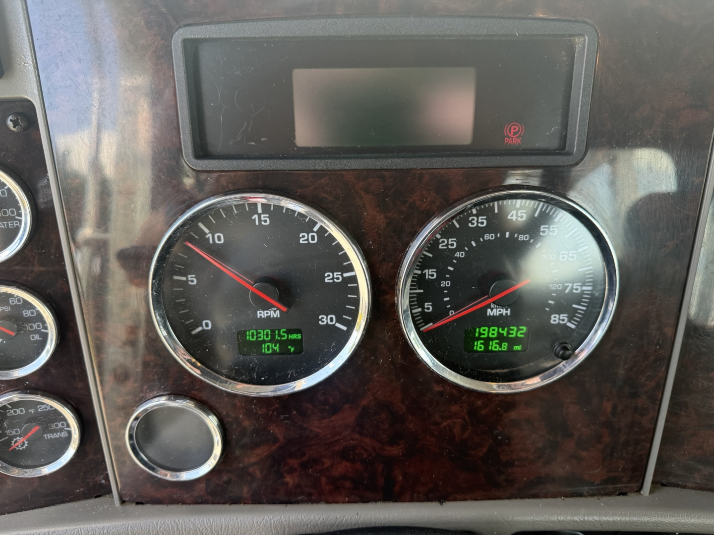 Kenworth truck instrument cluster and dashboard showing two round gauges with numbers, a small digital display above, and wood-grain panel. Left gauge marked RPM 0-30, right gauge shows speed/engine data, with digital readouts beneath.