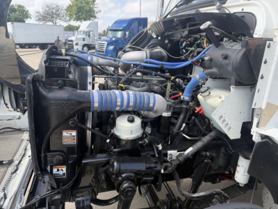 Truck hood open showing engine bay with blue hoses, black radiator housing, white reservoir, and various cables; Kenworth T270 engine area.