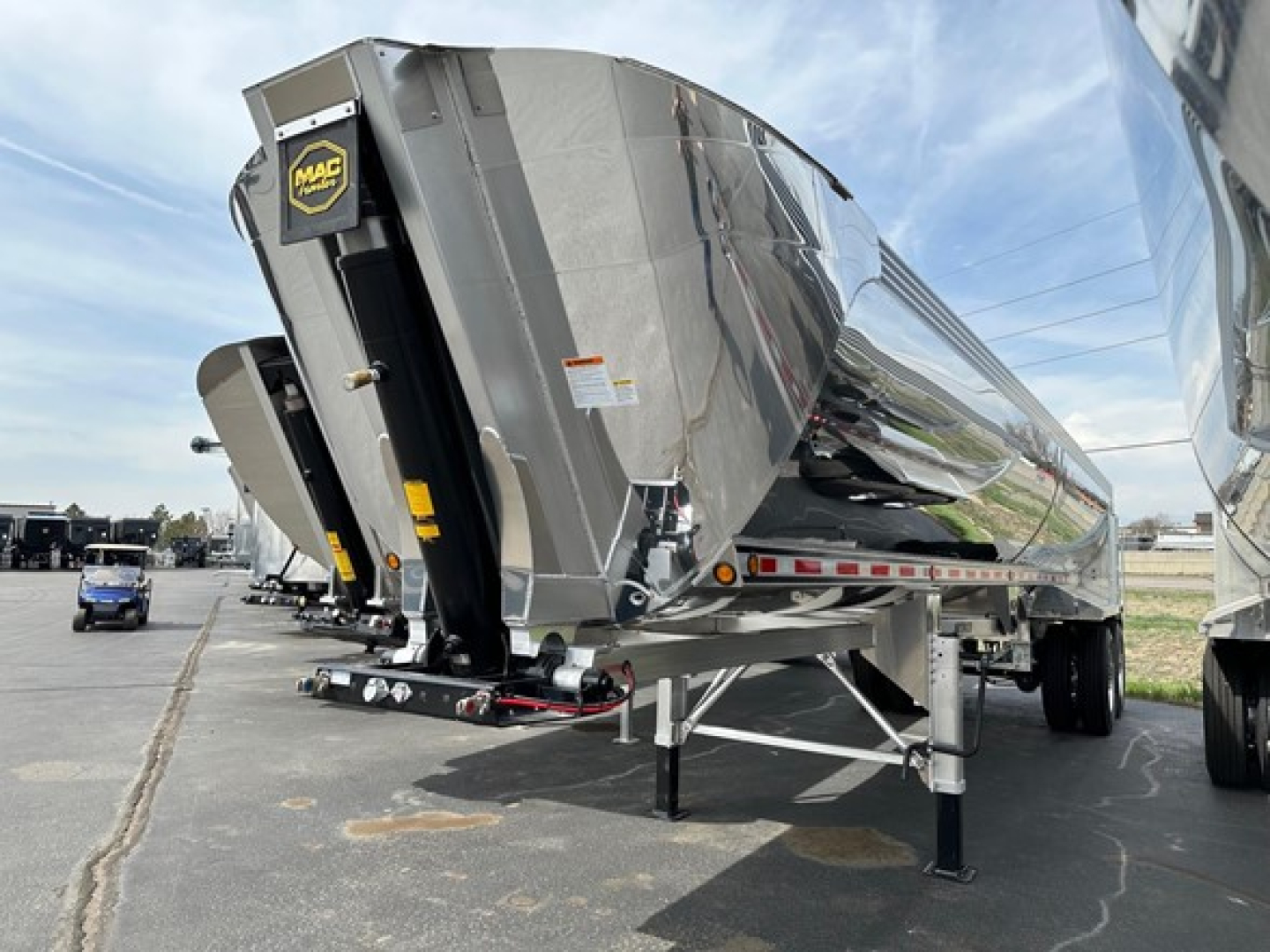 Dump trailer with MAC Macsimiser branding showing chrome metal hopper and support legs, rear with black access door, reflective tape, and front landing gear in a dealership lot.