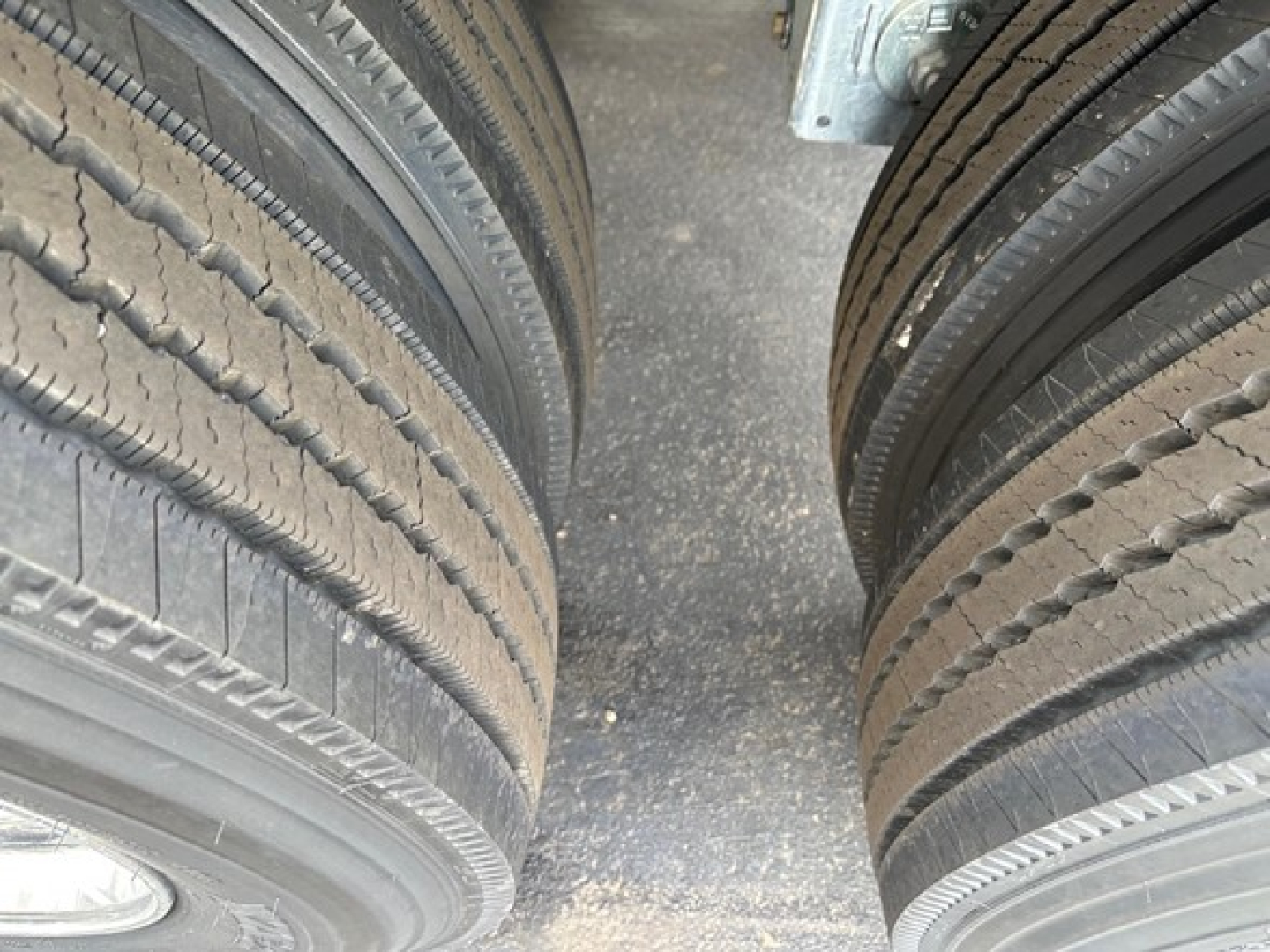 Dual truck tires mounted on a trailer with a textured asphalt surface between them. Visible tread patterns on both left and right tires.