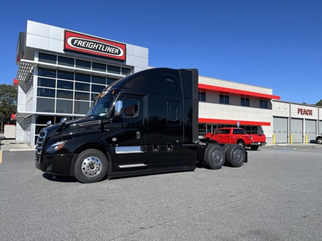 Freightliner Cascadia 126 tractor in black, front left view, with a sleek sleeper cab parked in front of a Freightliner dealership building.