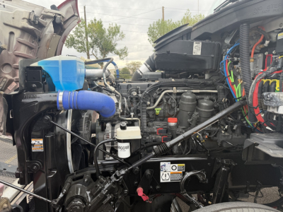 Kenworth T880 engine bay with visible fluid reservoirs and hoses, including blue coolant reservoir, white Rocar brake reservoir, and wiring loom.