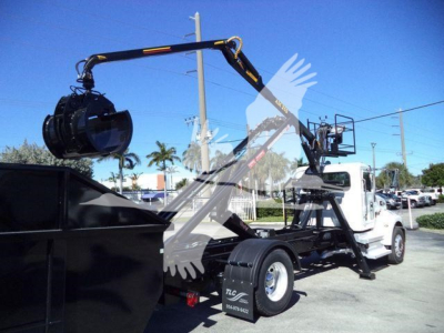 Peterbilt 337 roll-off truck with hydraulic arm and rotating hook lifting container, white cab, black chassis, in-service condition.