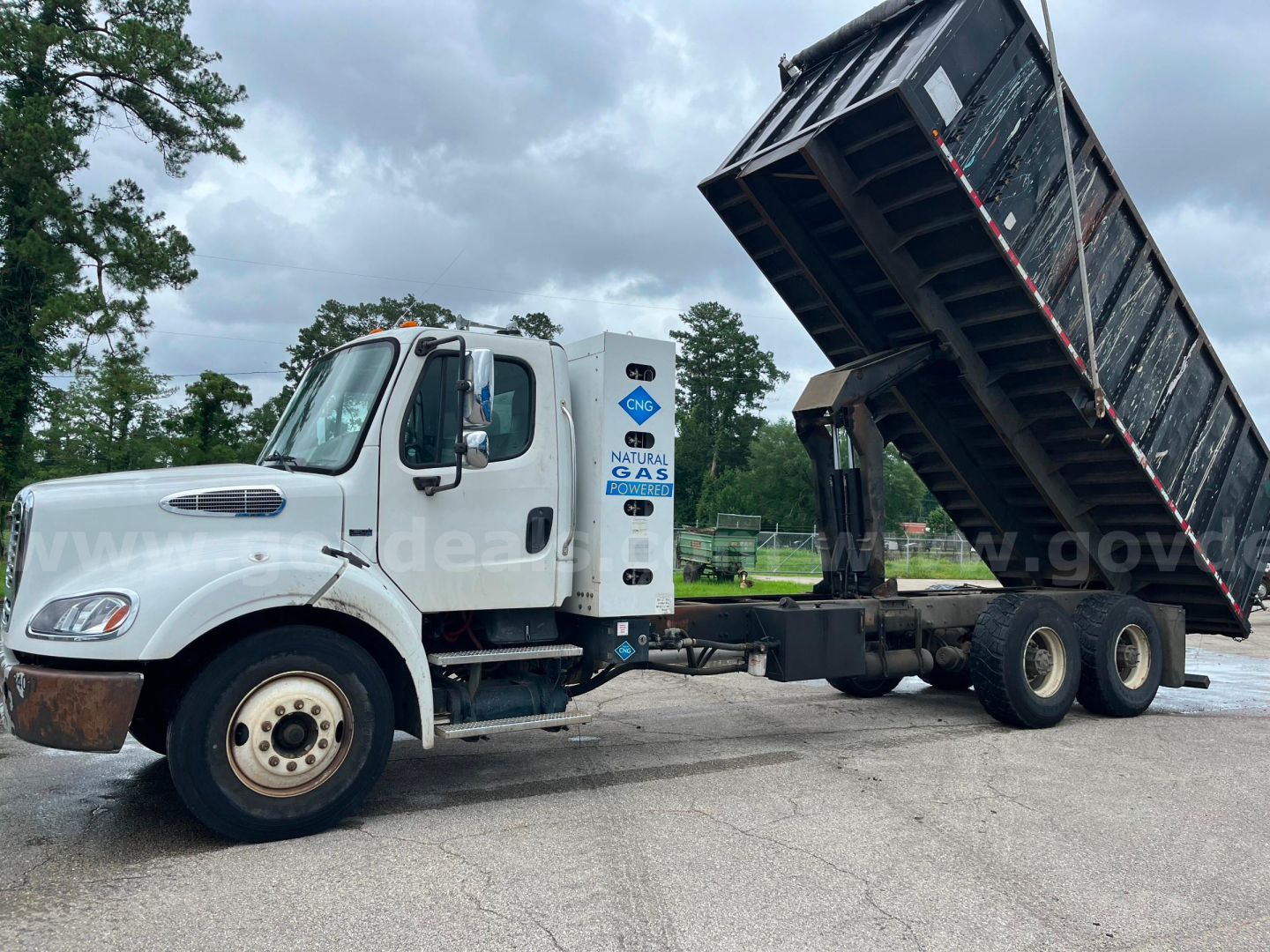 Dump truck in yard with raised black dump bed, white cab, and dual rear wheels. Freightliner M2 112 model with natural gas emblem on side and VIN tag present.