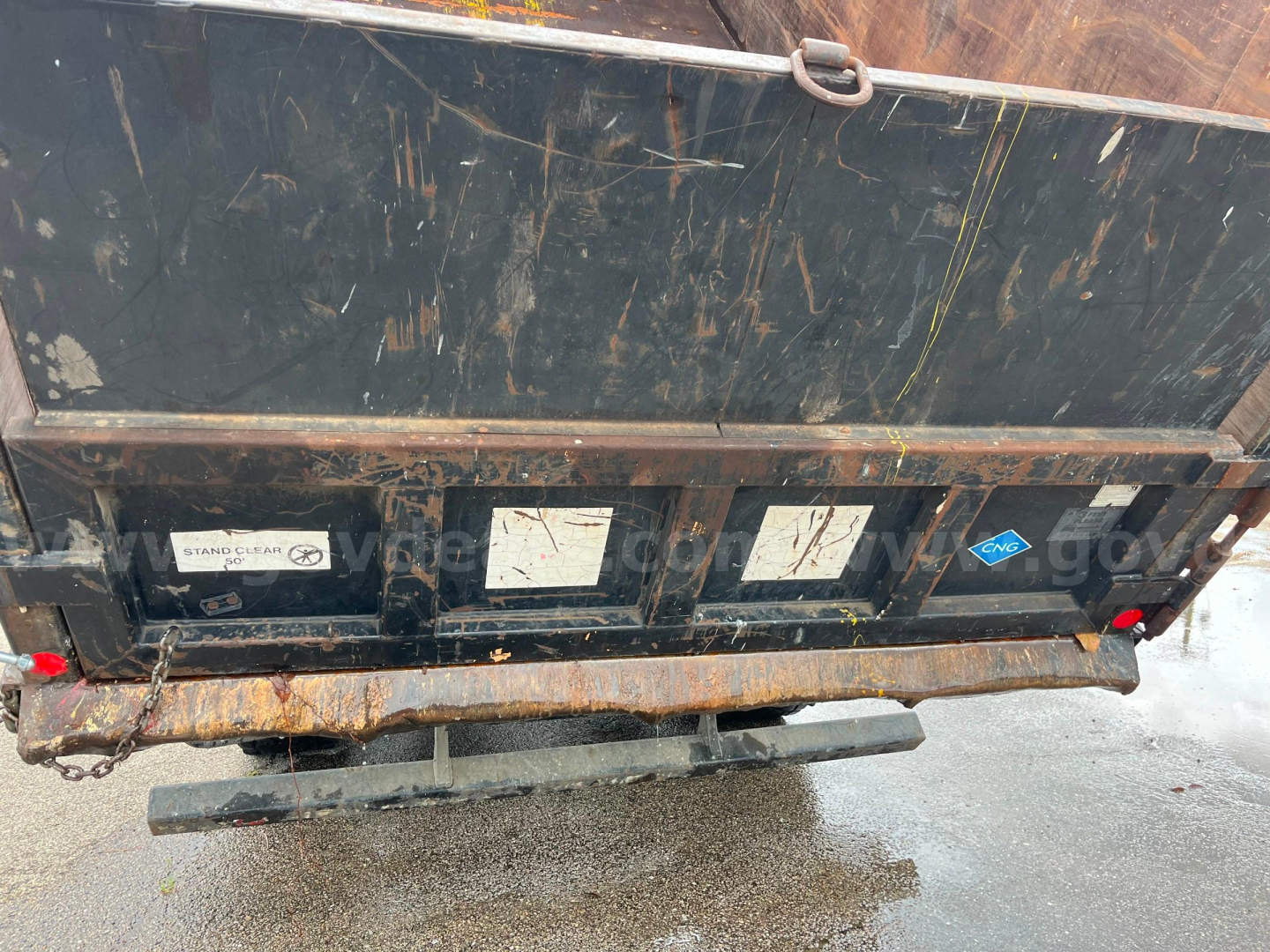 Truck bed of a Freightliner M2 112 dump with multiple stickers and a rusty bumper; visible labels include ‘Stand-...’ and a blue diamond sticker; wet ground beneath.