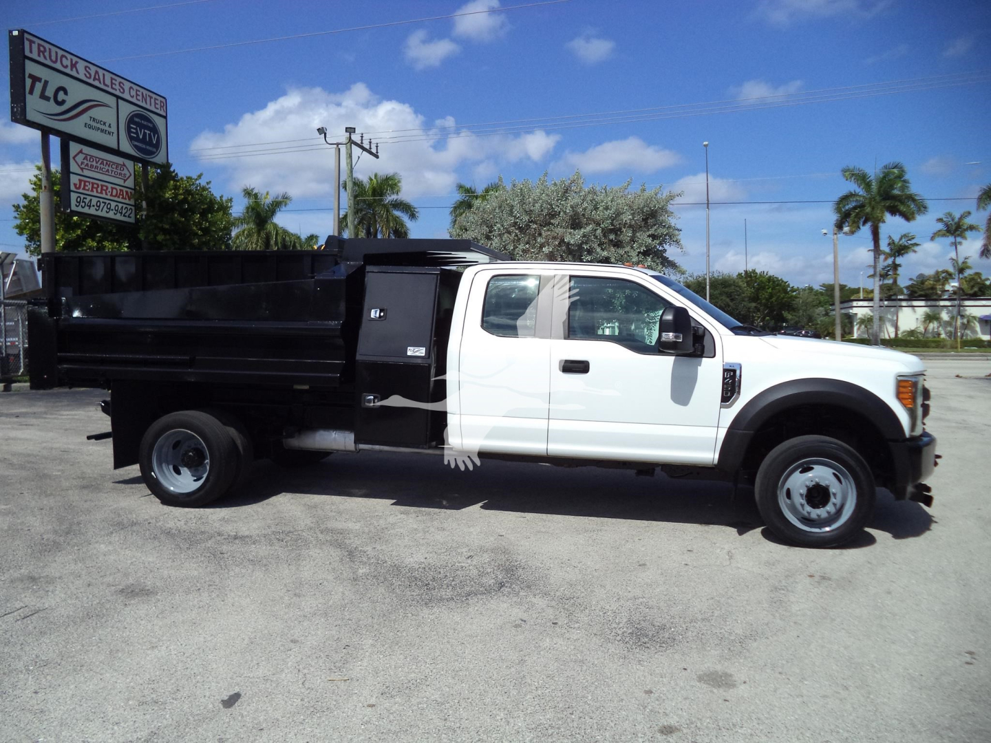 Ford F-450 dump truck side view showing white cab and black dump bed with TLC sign in background.