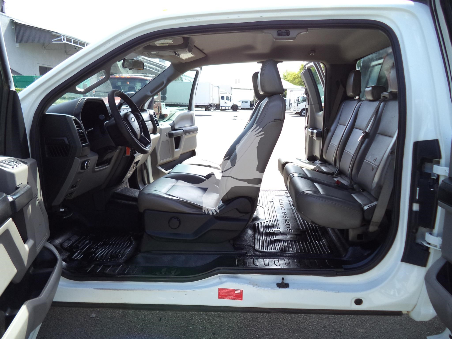 Ford F-450 XD interior showing front and rear seats with open passenger door and routing space, grey cloth seats, rubber floor mats, dashboard and steering wheel visible.