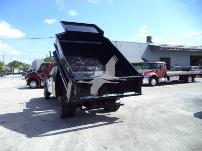 Dump truck with rear dump bed raised showing empty cargo space and wheels; no legible text on container. Scene includes other trucks in lot.
