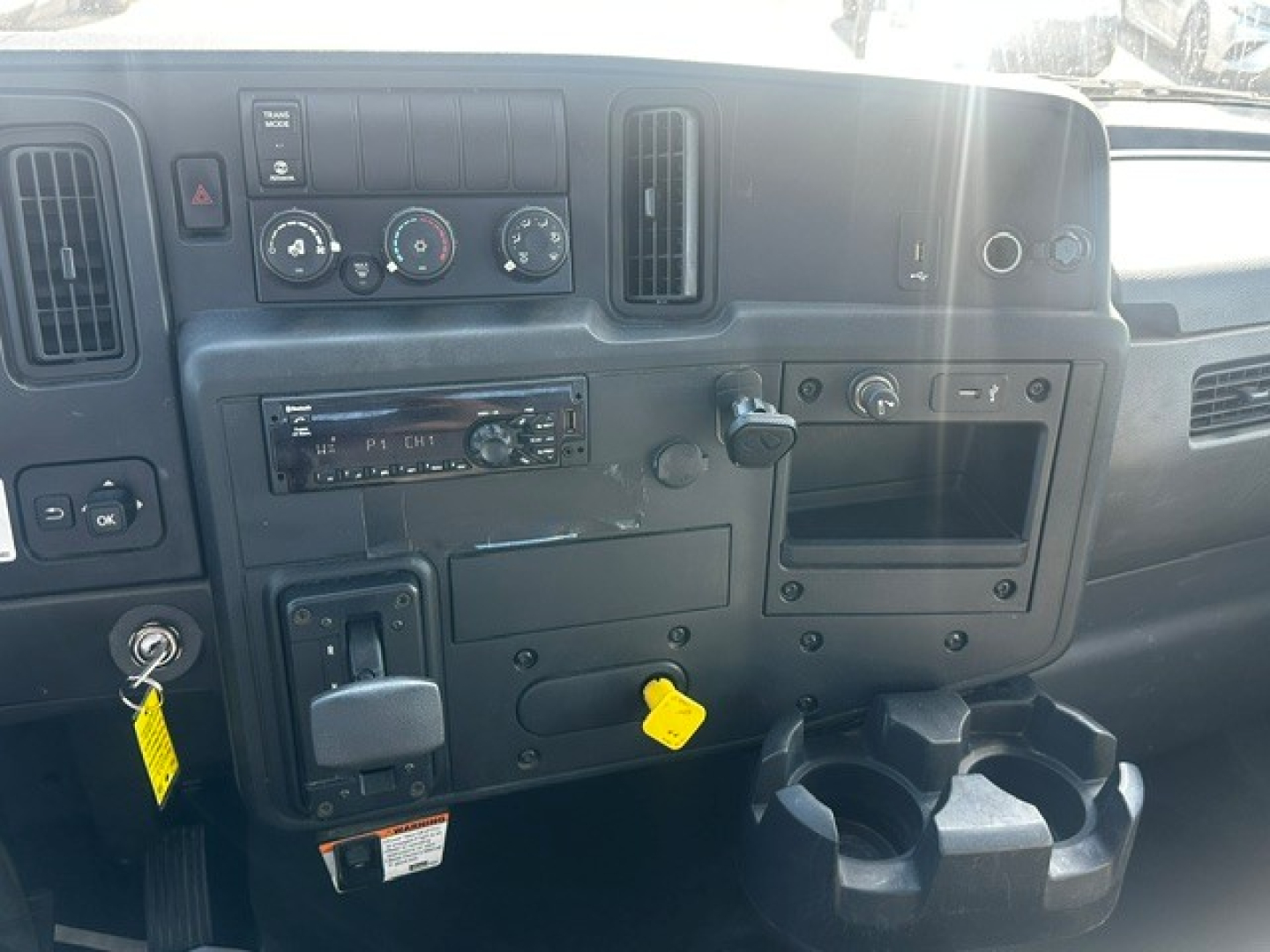 Truck dashboard interior showing ignition, radio, climate controls, cup holders, and yellow tag hanging from switch. Visible text includes keys and dashboard labels.