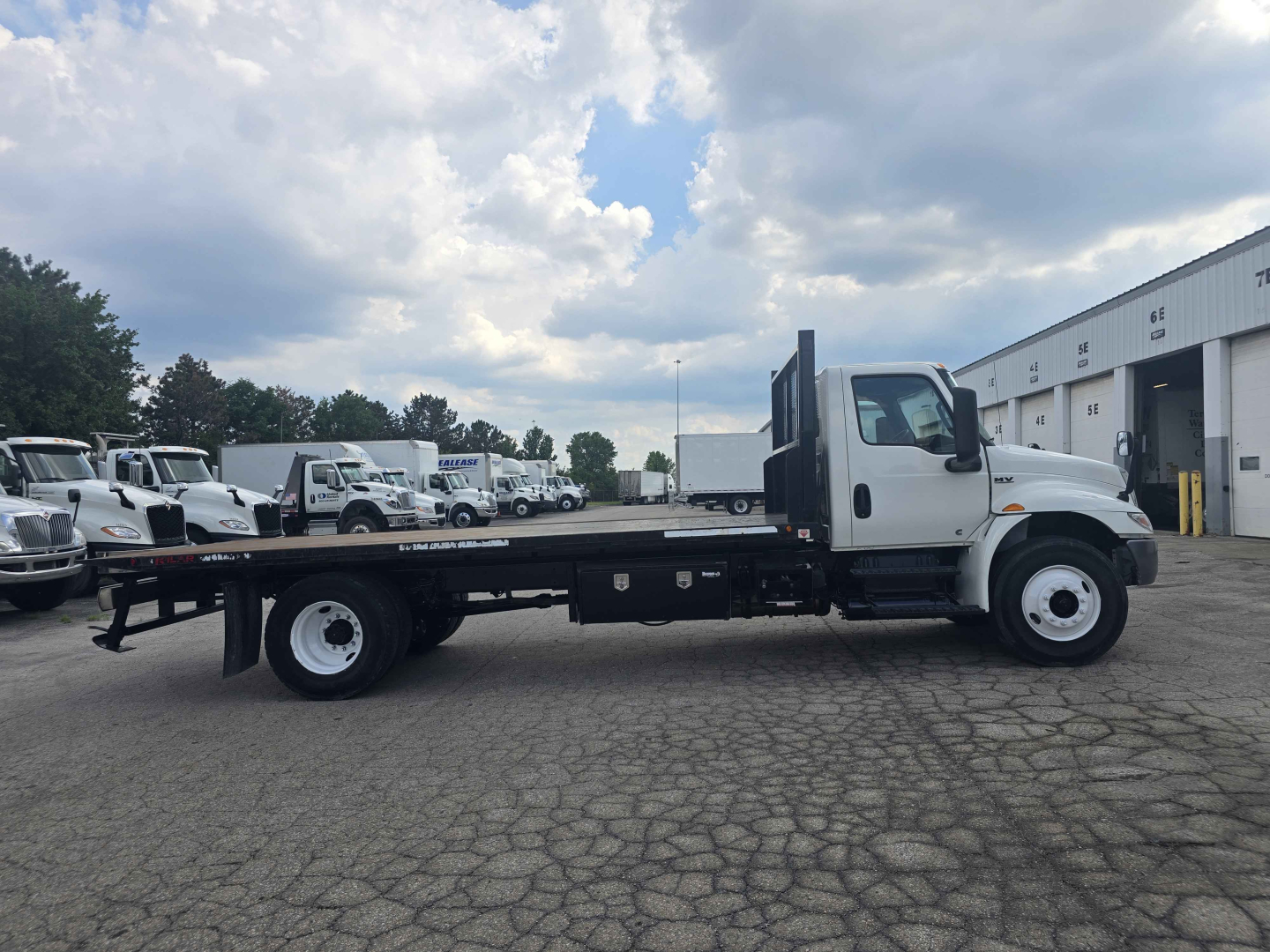 Flatbed rollback truck side view showing long black flatbed chassis, white cab, dual rear axles, and storage compartments under cab. No visible branding text legible.