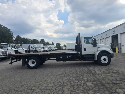 Flatbed rollback truck side view showing long black flatbed chassis, white cab, dual rear axles, and storage compartments under cab. No visible branding text legible.