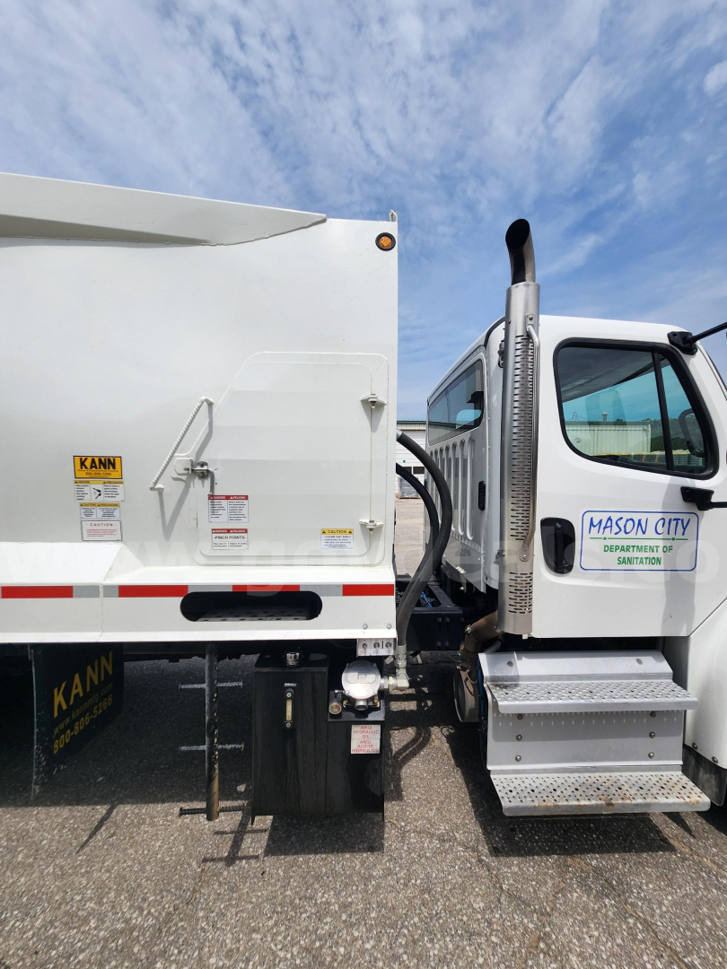 Truck side and cab of a Freightliner M2 106 with a garbage body. Visible text: 'KANN' on side steps, warning/sticker labels on rear dump, 'Mason City' on cab door, 'Deratti? sanitation' badge, hydraulic lines, steps, and rear lights.