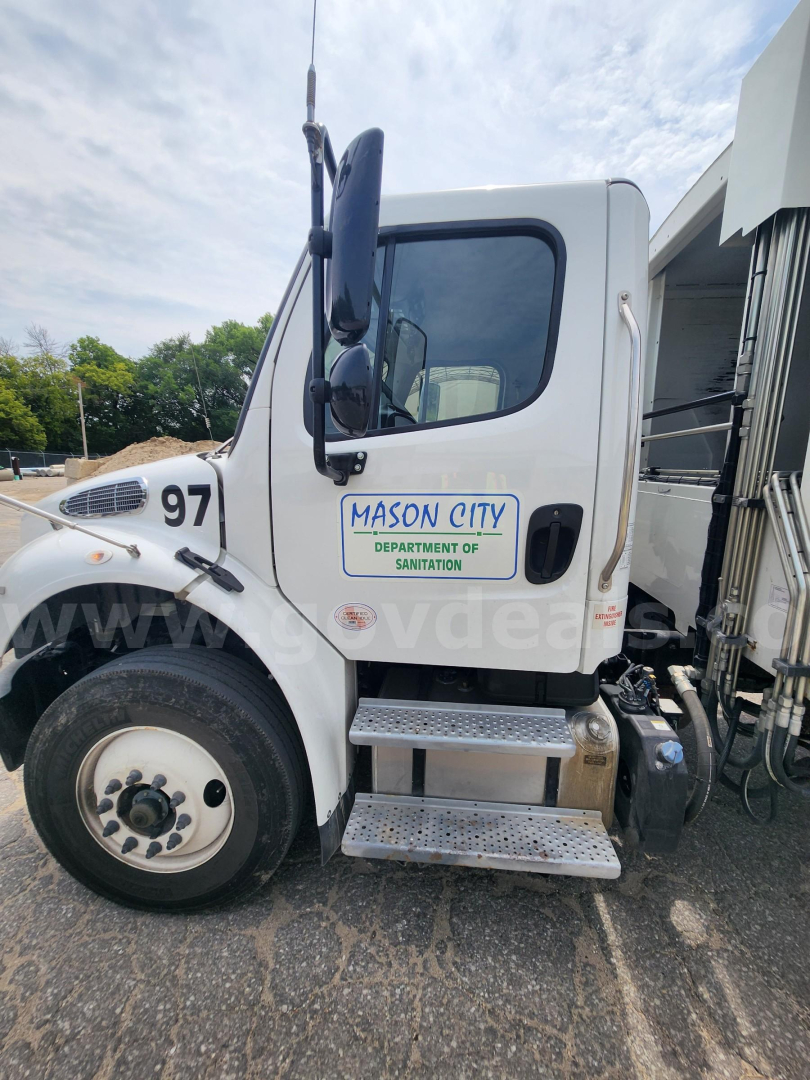 Truck cab side view showing white Freightliner M2 106 with Mason City Department of Sanitation logo on door, number 97 on fender, metal steps, and side mirror.