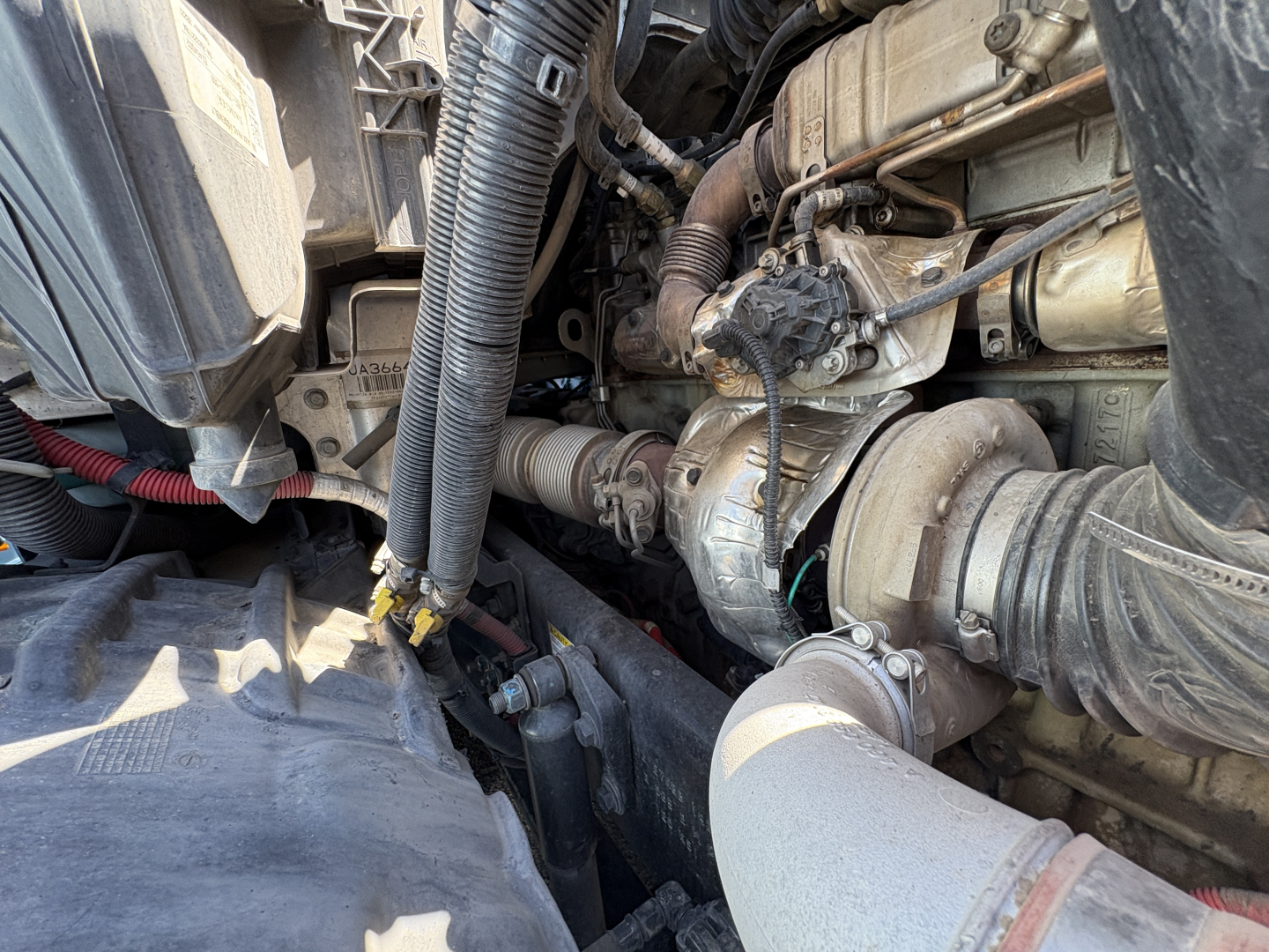 Truck engine bay view showing hoses, turbo, and intake on a Freightliner Cascadia 126 engine. Visible components include air intake hose, turbocharger, coolant lines, and wiring harnesses.