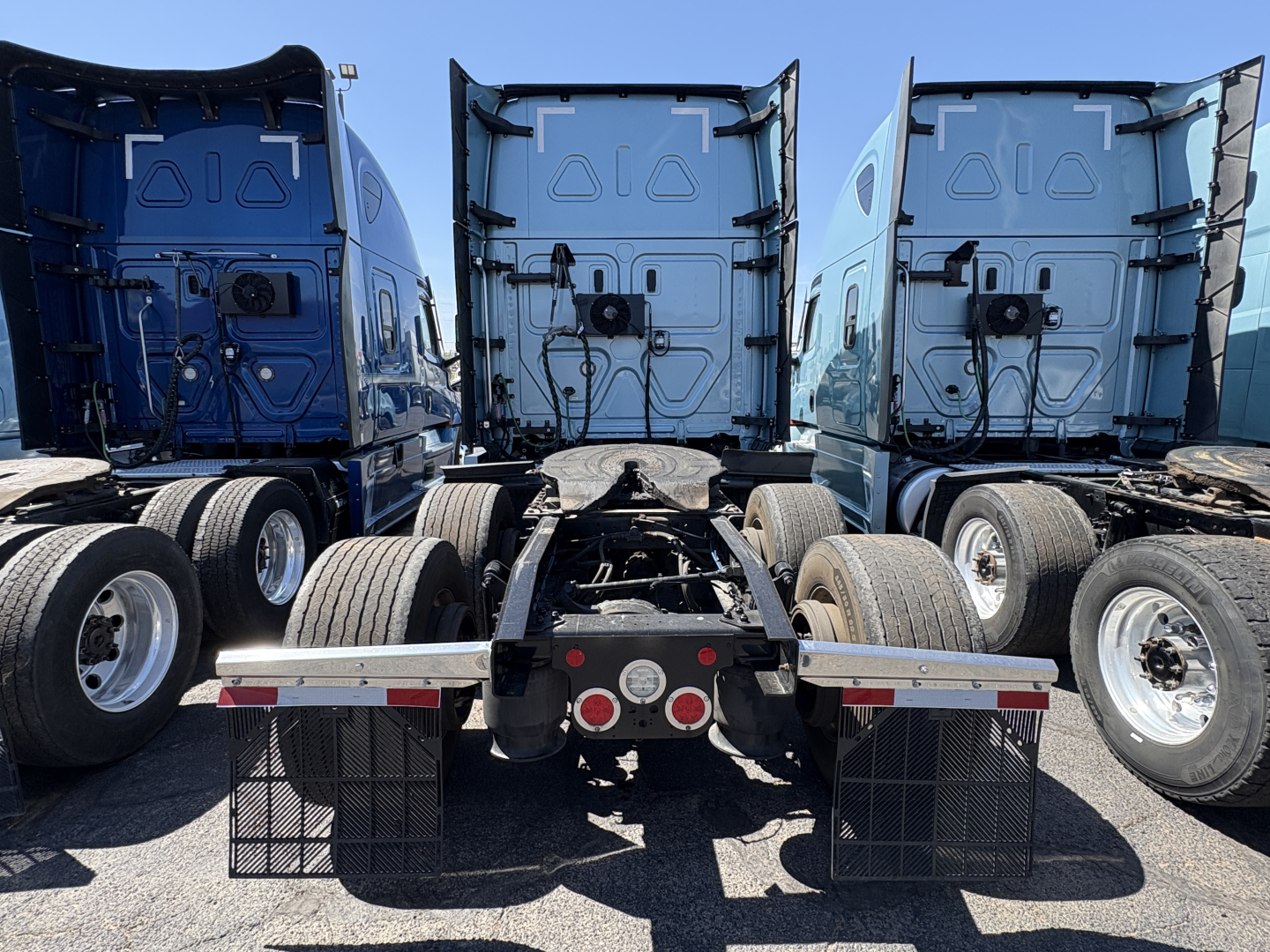Rear view of a Freightliner Cascadia 126 semi truck tractor with fifth wheel and dual rear axles, three blue sleeper cabs in a row, visible mudflaps and rear lights.